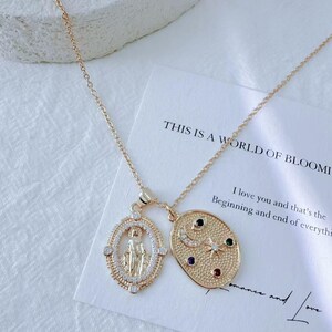 May include: Gold-toned necklace with two pendants. One pendant is an oval with a religious figure, the other is an oval with a moon, stars, and colorful gems. The necklace is on a white card with text.