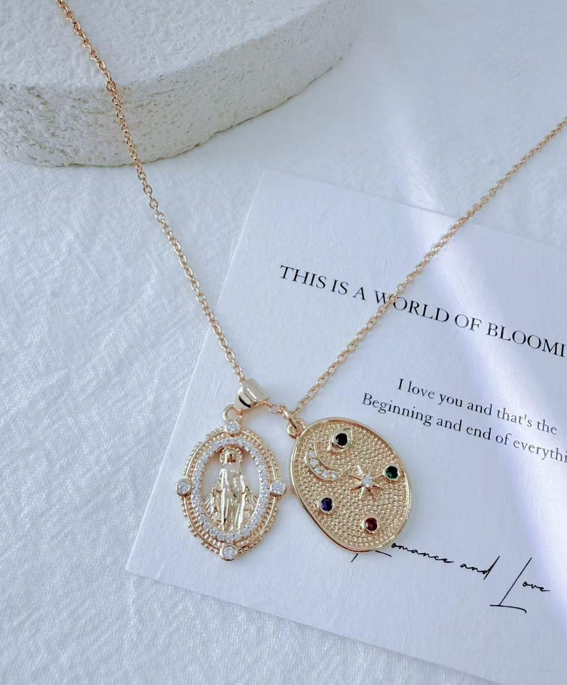 May include: Gold-toned necklace with two pendants. One pendant is an oval with a religious figure, the other is an oval with a moon, stars, and colorful gems. The necklace is on a white card with text.