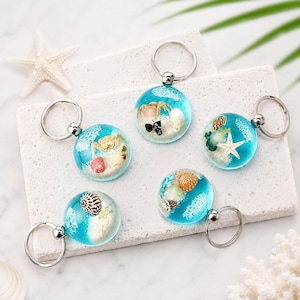 May include: Five circular keychains featuring clear blue resin domes filled with miniature seashells, sand, and small crab figures. Each keychain has a silver-toned metal ring attached to the top, showcasing a beach-themed design with white coral-like patterns embedded within the resin.