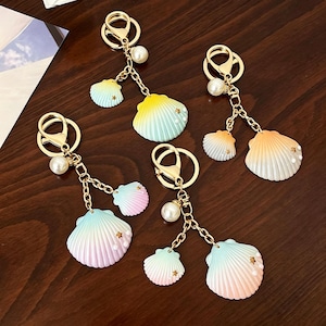 May include: Four gold-toned metal keychains featuring gradient-coloured seashell charms. Each keychain includes a lobster clasp, a round ring, a single white faux pearl bead, and two scalloped shell pendants decorated with tiny gold stars and pearls.
