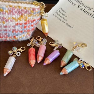 May include: A collection of colorful pencil-shaped charms with gold-tone hardware. The charms are in various colors, including white, red, pink, purple, green, and blue. A small, patterned pouch is visible in the background.