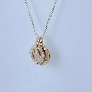 May include: Gold-toned pendant necklace featuring a detailed religious figure within a circular frame. The pendant is adorned with small, sparkling accents and a delicate chain. The necklace is displayed against a plain white background.