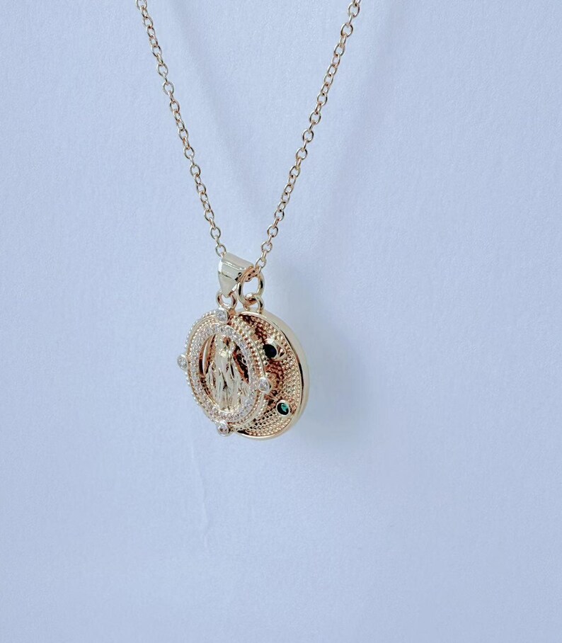 May include: Gold-toned pendant necklace featuring a detailed religious figure within a circular frame. The pendant is adorned with small, sparkling accents and a delicate chain. The necklace is displayed against a plain white background.