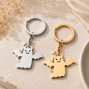 Könnte beinhalten: Zwei Schlüsselanhänger mit Halloween-Motiv. Einer ist silberfarben, der andere goldfarben. Jeder Schlüsselanhänger zeigt ein teuflisches Geisterdesign mit Flügeln, einer Mistgabel und einem lächelnden Gesicht. Die Schlüsselanhänger sind an einem Metallring und einer Kette befestigt.