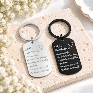 May include: Two rectangular metal keychains, one silver and one black, each featuring a split ring. Both are engraved with the text "Little Reminders:" followed by five positive affirmations and a heart icon, including "You are enough" and "Be kinder to yourself."