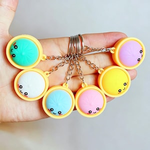 May include: A collection of colorful, round keychains with smiling faces. Each keychain features a different pastel color, including green, white, blue, pink, yellow, and purple, set within a yellow frame. The keychains are attached to silver key rings.