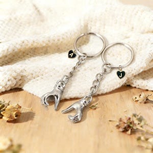 May include: Two silver keyrings with hand-shaped charms. Each keyring has a heart-shaped charm with a letter, one with "A" and the other with "J". The keyrings are on a white knitted surface.