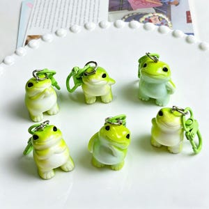 May include: A collection of six frog-shaped keychains. Each keychain features a small, rounded frog figurine in shades of green and white, with a silver-colored metal ring and a green clasp. The frogs have black eyes and are arranged on a white plate.