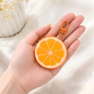 May include: A circular orange slice keychain featuring a realistic citrus fruit design with vibrant orange segments and a white rind. The keychain is attached to a small, bright orange plastic lobster clasp connector for easy attachment to items.