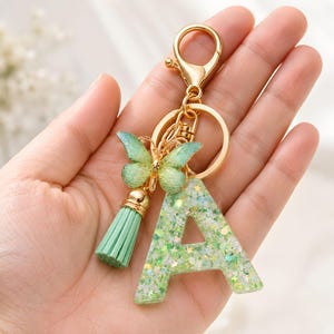 Può includere: Un portachiavi in metallo dorato con un ciondolo a forma di lettera A in resina verde chiaro con glitter e stelline incorporate. All'anello sono fissati un piccolo ciondolo a farfalla verde chiaro testurizzato e una nappa verde chiaro.