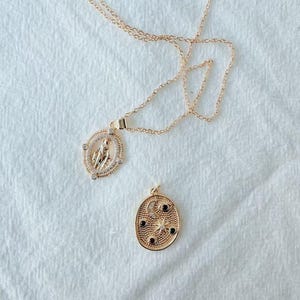 May include: Two gold-colored necklaces on a white surface. One necklace features a pendant with a religious figure inside a circular frame with clear stones. The other necklace has a pendant with a moon, stars, and black stones.