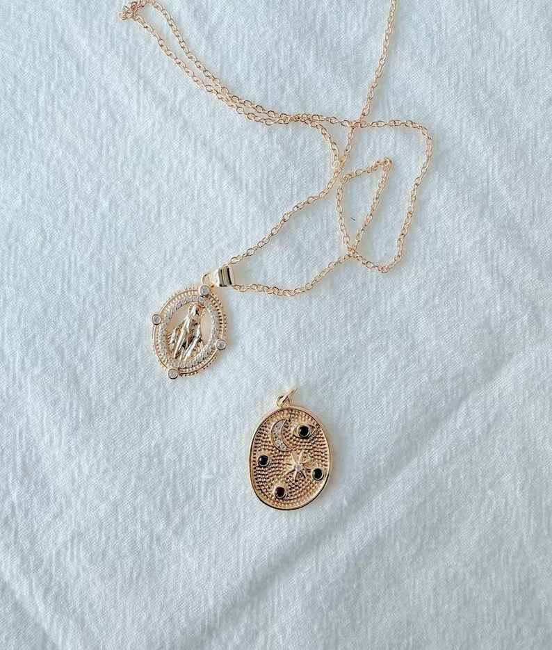 May include: Two gold-colored necklaces on a white surface. One necklace features a pendant with a religious figure inside a circular frame with clear stones. The other necklace has a pendant with a moon, stars, and black stones.