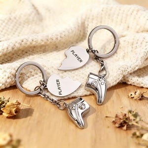 Peut inclure: Deux porte-clés argentés, chacun avec un motif de cœur brisé. Une moitié du cœur porte l'inscription "PLAYER" et est attachée à un charm en forme de manette. Les porte-clés sont sur une surface en bois avec une couverture en tricot crème en arrière-plan.