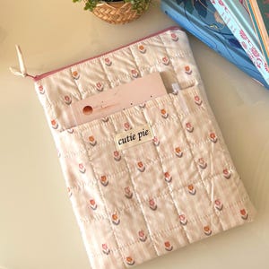 Puede incluir: Una funda acolchada para tablet con un estampado floral en blanco roto y rosa, con cremallera y bolsillo rosas. La etiqueta dice "cutie pie". Una pequeña planta en maceta y dos libros están en el fondo.