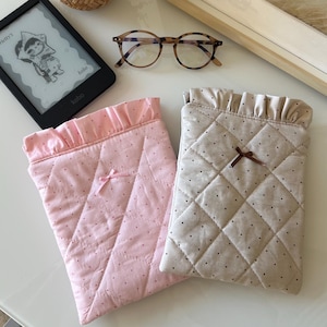 Könnte beinhalten: Zwei rechteckige, gesteppte Taschen, eine in Hellrosa und eine in Beige, beide mit einem kleinen gerüschten oberen Rand, einer winzigen mittigen Schleife und einem dezenten Punktemuster. Die Taschen liegen neben einem schwarzen E-Reader und einer Brille.