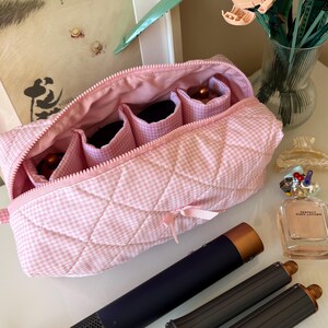 Könnte beinhalten: Eine rosa-weiß karierte, gesteppte Kosmetiktasche mit Reißverschluss, die Fächer enthält. Ein dunkelblaues Haarstyling-Tool mit zwei schwarzen Aufsätzen und einem goldenen Akzent befindet sich neben der Tasche. Ein gerahmtes Bild und eine Vase mit Blumen sind im Hintergrund.