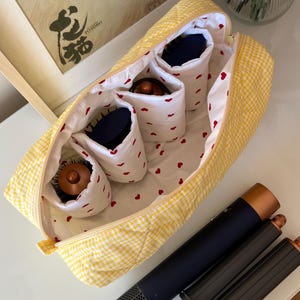 May include: A yellow gingham cosmetic bag with white interior and red heart pattern. The bag contains several hairbrushes and a black and copper hair styling tool. A framed picture is in the background.
