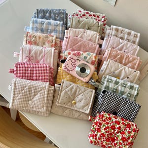 May include: A collection of small, quilted fabric pouches in various patterns including floral, gingham, polka dot, and cherry prints, featuring snap closures. A pink Hello Kitty digital camera is placed among the pouches on a white surface.