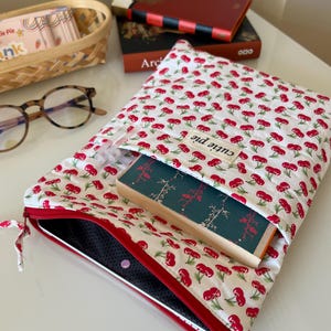 以下が含まれることがあります： 赤いサクランボ柄の白いブックカバー。赤いジッパーと本が入っています。カバーには「Cutie Pie」と書かれた小さなタグが付いています。他の本とバスケットが背景にあります。