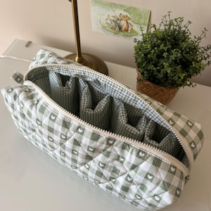 May include: A rectangular quilted storage pouch featuring a sage green and white gingham pattern with small white heart icons. The pouch has a white zipper closure and an interior divided into multiple vertical compartments with matching fabric.