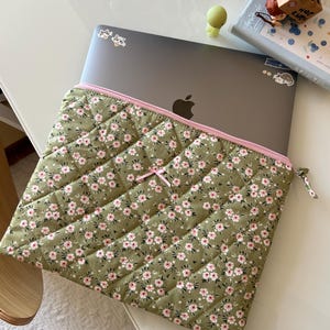 Peut inclure: Une housse d'ordinateur portable matelassée vert olive avec un petit motif floral rose et blanc. La housse présente une fermeture éclair rose clair, un petit nœud en ruban rose au centre et contient un ordinateur portable gris foncé avec un logo Apple.