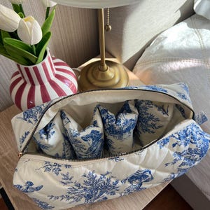 May include: A quilted toiletry bag with a blue and white pastoral design. The bag is open, revealing several interior compartments. A gold zipper and white interior lining complete the design. The bag is on a wooden surface.