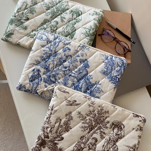 Può includere: Tre trousse per cosmetici trapuntate con motivi toile de Jouy in verde, blu e marrone. Ogni borsa ha un motivo a rombi e una chiusura con cerniera. Sono visibili anche un taccuino, una penna e degli occhiali.