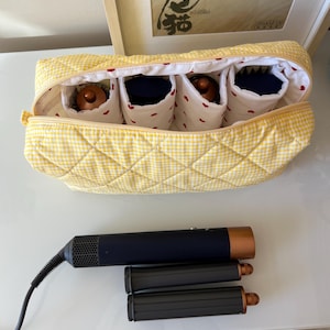 May include: A yellow gingham quilted cosmetic bag with four interior compartments, each holding a hair styling tool. Below, a black and copper hair styling tool and two black attachments rest on a white surface.