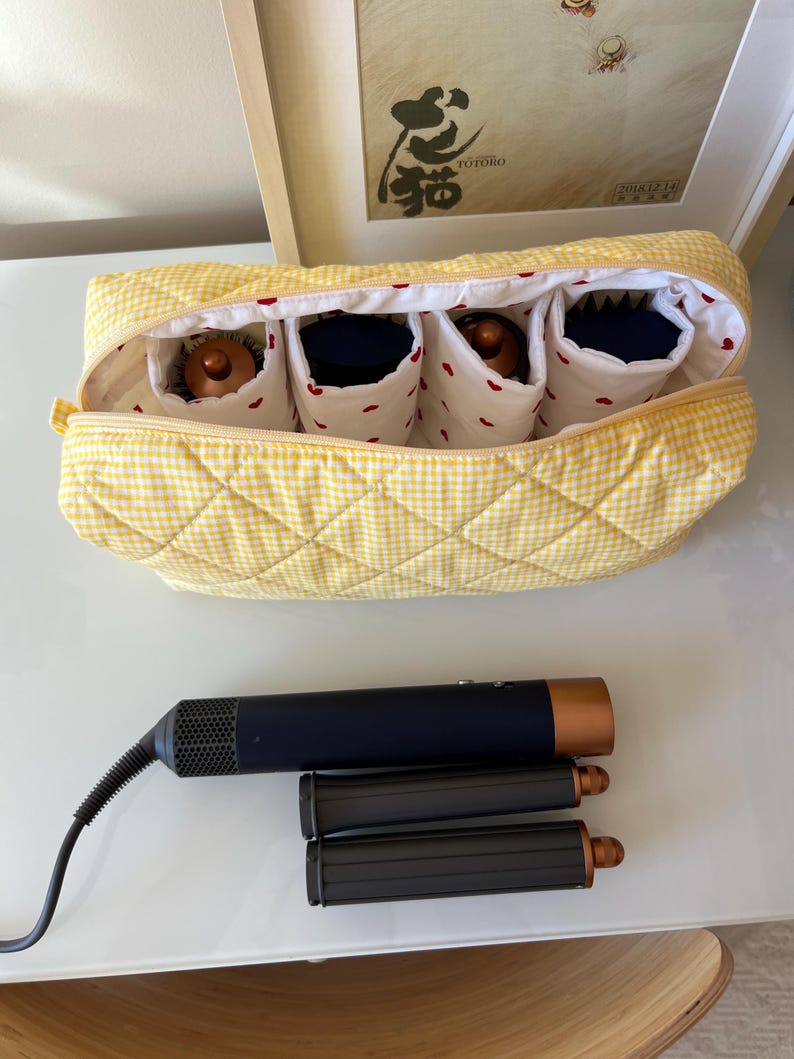 May include: A yellow gingham quilted cosmetic bag with four interior compartments, each holding a hair styling tool. Below, a black and copper hair styling tool and two black attachments rest on a white surface.