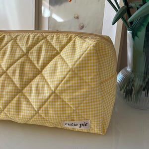 May include: A yellow and white gingham quilted cosmetic bag with a zipper closure. The bag has a rectangular shape and features the text "cutie pie" on a small label.