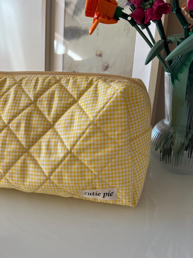 May include: A yellow and white gingham quilted cosmetic bag with a zipper closure. The bag has a rectangular shape and features the text "cutie pie" on a small label.