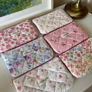 Puede incluir: Varias bolsas de cosméticos acolchadas con diseños florales en tonos rosa, blanco y azul. Cada bolsa tiene una cremallera dorada y una etiqueta que dice "cutie pie".