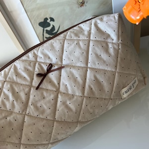 Pu&ograve; includere: Un beauty case trapuntato beige con cerniera marrone e un piccolo fiocco marrone. La borsa &egrave; decorata con piccoli pois neri e ha un'etichetta con la scritta "cutie pie".