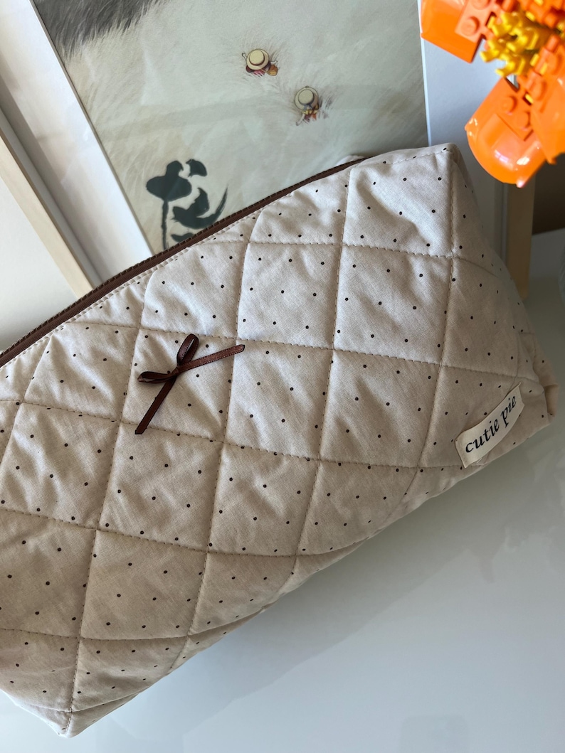 Pu&ograve; includere: Un beauty case trapuntato beige con cerniera marrone e un piccolo fiocco marrone. La borsa &egrave; decorata con piccoli pois neri e ha un'etichetta con la scritta "cutie pie".