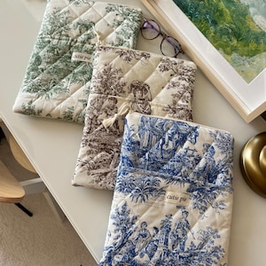 Può includere: Quattro custodie in tessuto trapuntato verde, marrone e blu con motivi toile. Ogni custodia ha una chiusura a cerniera e una piccola etichetta con la scritta "cutie pie". Le custodie sono disposte su una superficie bianca.