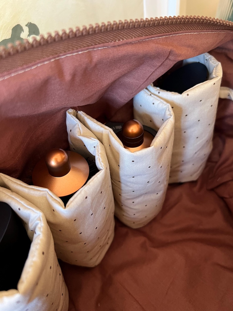Pu&ograve; includere: Vista aperta di una borsa da viaggio marrone con cerniera. All'interno, ci sono diverse tasche in tessuto color crema con piccoli pois neri. Le tasche contengono oggetti color rame. L'interno della borsa &egrave; marrone rossiccio.