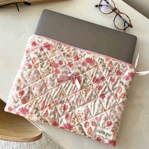 Può includere: Una custodia trapuntata per laptop con un motivo floreale rosa, pesca e verde su sfondo crema. La custodia ha un bordo rosa e un piccolo fiocco. Il testo "cutie pie" è su una piccola etichetta.