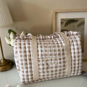 May include: A quilted tote bag featuring a beige and white gingham pattern with a ruffled top edge. The bag has two beige fabric handles and a front pocket with a small white label that reads cutie pie. A detachable beige shoulder strap is included.