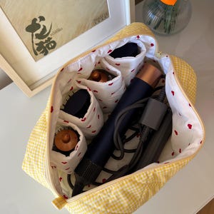 May include: A yellow gingham cosmetic bag with a white interior patterned with red hearts. The bag is open, revealing compartments holding hair styling tools. A framed picture and a vase with orange flowers are in the background.