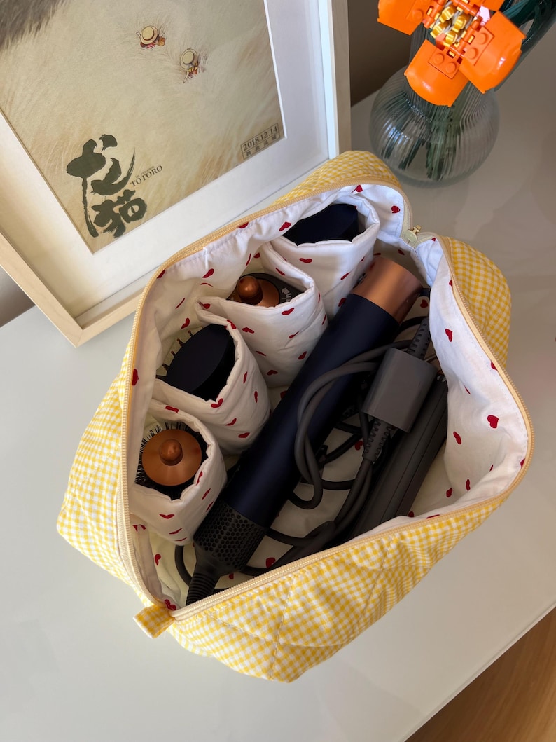 May include: A yellow gingham cosmetic bag with a white interior patterned with red hearts. The bag is open, revealing compartments holding hair styling tools. A framed picture and a vase with orange flowers are in the background.