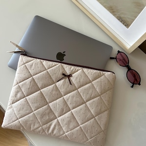 May include: A beige quilted laptop sleeve with a burgundy zipper and bow detail. The sleeve is on a white surface next to a silver laptop, sunglasses, and a framed picture.