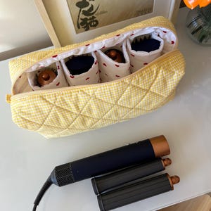 May include: A yellow gingham quilted cosmetic bag with four interior compartments, each holding a beauty product. A black and copper hair styling tool and two attachments are next to the bag.