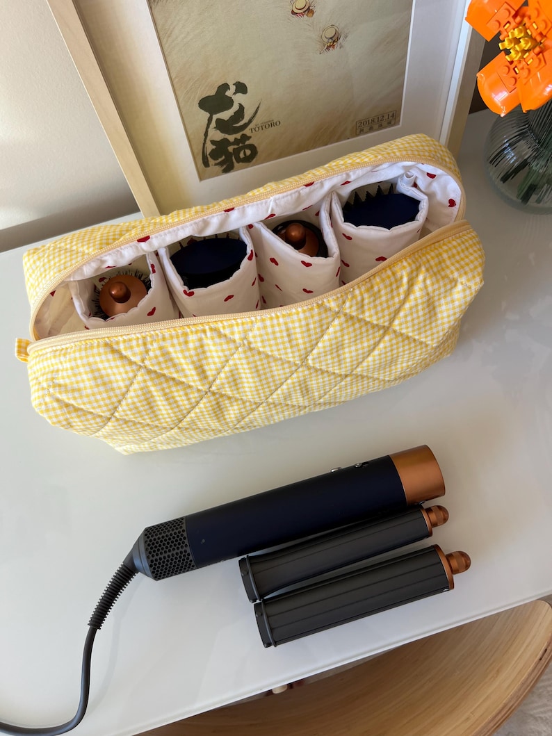 May include: A yellow gingham quilted cosmetic bag with four interior compartments, each holding a beauty product. A black and copper hair styling tool and two attachments are next to the bag.
