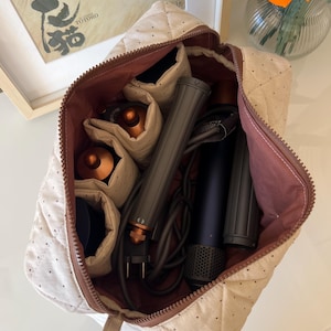 Pu&ograve; includere: Un beauty case trapuntato beige con cerniera e interno marrone, aperto per rivelare strumenti per lo styling dei capelli. La borsa contiene un asciugacapelli, un arricciacapelli e altri accessori. La borsa &egrave; su una superficie bianca.