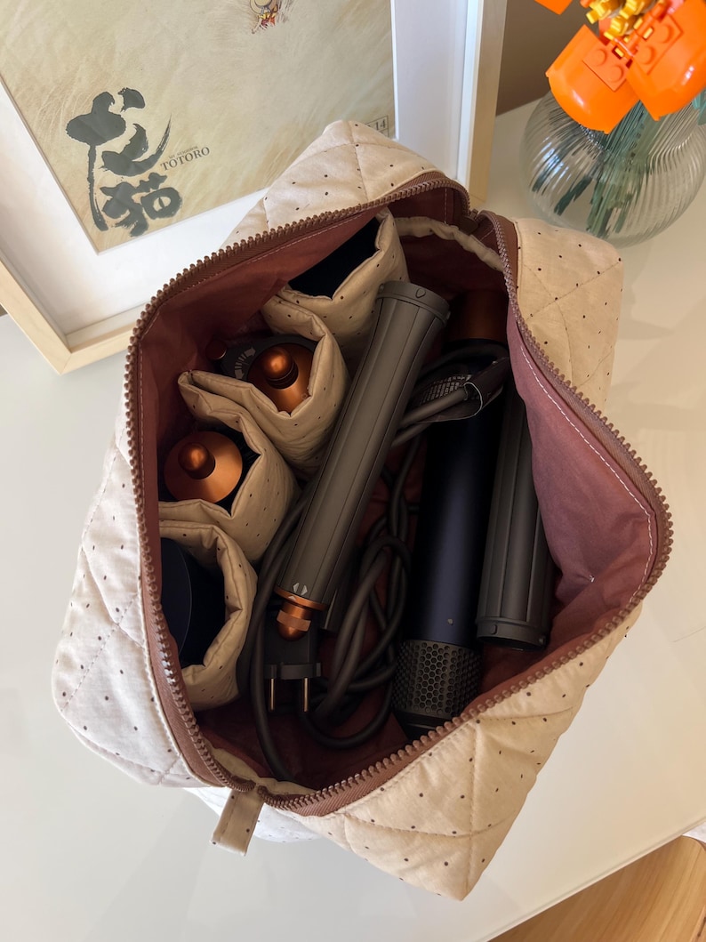 Pu&ograve; includere: Un beauty case trapuntato beige con cerniera e interno marrone, aperto per rivelare strumenti per lo styling dei capelli. La borsa contiene un asciugacapelli, un arricciacapelli e altri accessori. La borsa &egrave; su una superficie bianca.
