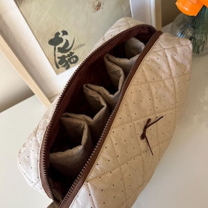 Pu&ograve; includere: Borsa per cosmetici trapuntata beige con pois marroni e cerniera marrone. L'interno presenta pi&ugrave; scomparti. Un piccolo fiocco marrone &egrave; attaccato all'esterno. La borsa &egrave; aperta, rivelando l'interno.