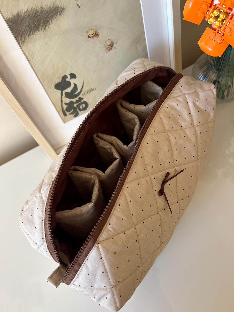 Pu&ograve; includere: Borsa per cosmetici trapuntata beige con pois marroni e cerniera marrone. L'interno presenta pi&ugrave; scomparti. Un piccolo fiocco marrone &egrave; attaccato all'esterno. La borsa &egrave; aperta, rivelando l'interno.