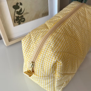 May include: A yellow and white gingham patterned cosmetic bag with a gold zipper. The rectangular bag is quilted and has a gold zipper pull. The bag is sitting on a white surface.