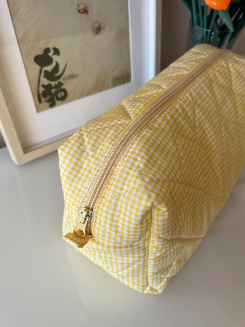 May include: A yellow and white gingham patterned cosmetic bag with a gold zipper. The rectangular bag is quilted and has a gold zipper pull. The bag is sitting on a white surface.