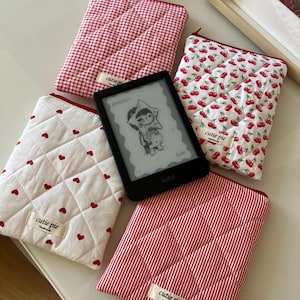 Può includere: Quattro custodie in tessuto trapuntato con motivi rossi e bianchi, tra cui quadretti vichy, stampa a ciliegie, piccoli cuori e righe verticali. Ogni custodia presenta un'etichetta beige con la scritta cutie pie handmade e un e-reader Kobo nero.
